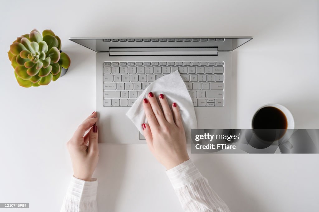 Eine Arbeiterin, die mit Antiviren-Nass putzt, wischt einen Laptop und einen Schreibtisch im Büro, bevor sie mit der Arbeit beginnt, um sich vor Bakterien und Viren zu schützen.