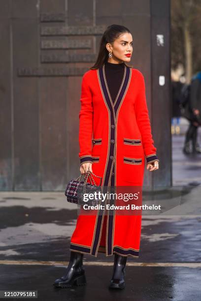 Camila Coelho wears earrings, a black turtleneck pullover, a red long wool jacket with buttons and pockets, a tweed checked colored Chanel bag, black...