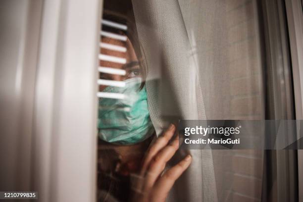 young woman in quarantine wearing a mask and looking through the window - quarantine stock pictures, royalty-free photos & images