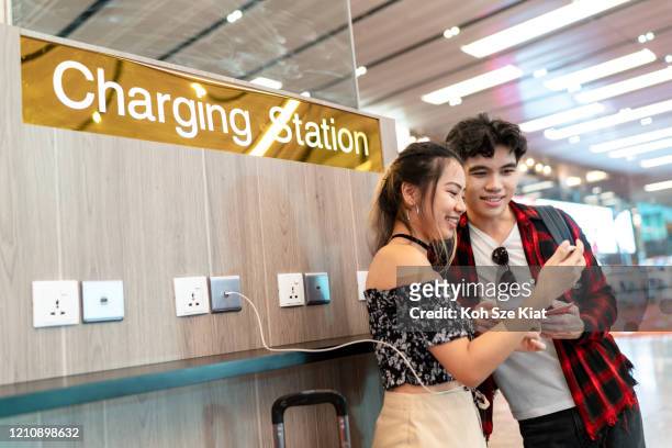 joven pareja asiática disfrutando de uso gratuito de por teléfono en el aeropuerto - estación de carga eléctrica fotografías e imágenes de stock