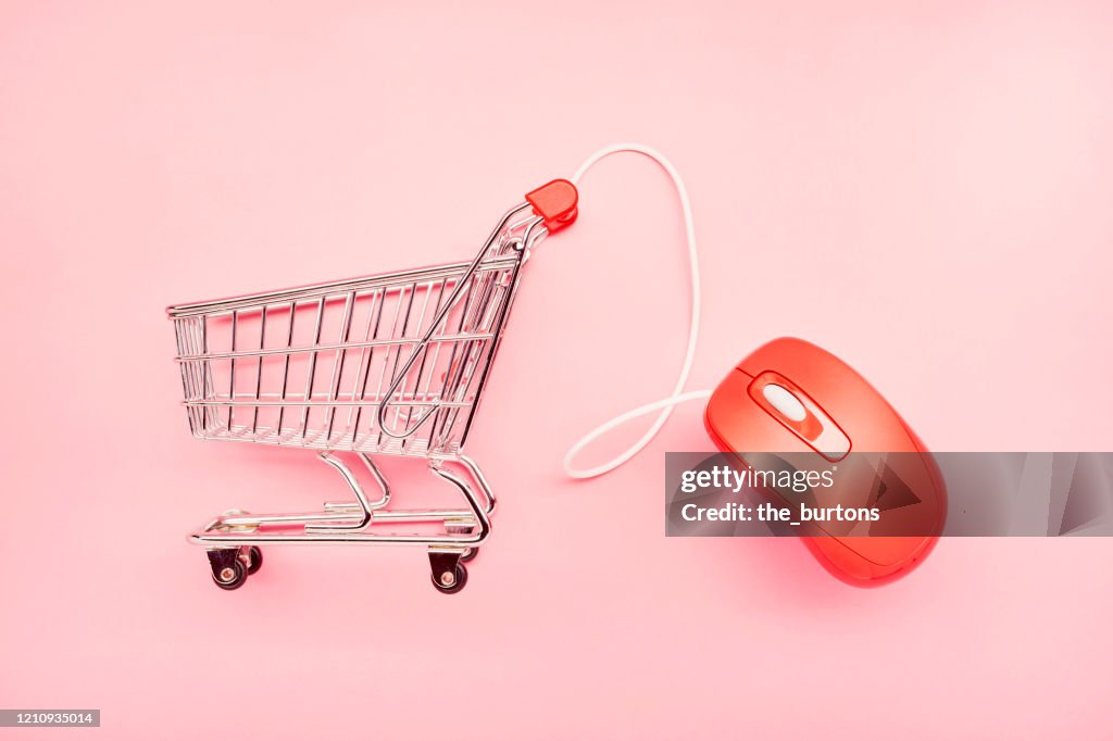 Still life of a small shopping cart and red computer mouse on pink background, online shopping