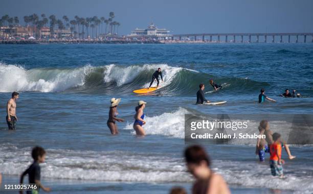 Surfer catches a wave while joining some of the thousands of beach-goers enjoying a warm, sunny day at the beach amid state-mandated stay-at-home and...