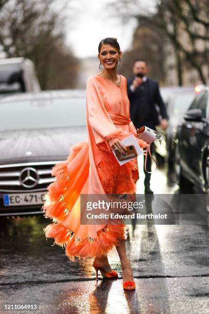 Helena Bordon wears an orange lustrous and silky dress with fluffy parts, earrings, a bag, orange high heels shoes outside Valentino, during Paris...