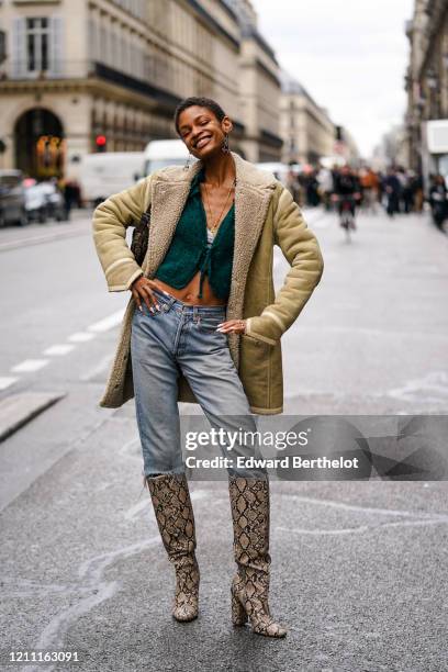 Model wears earrings, a necklace, a green knit crop top with a plunging neckline, ripped blue jeans, a beige shearling jacket, light brown and black...