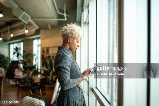 mujer de empresa tatuada usando una tableta digital cerca de la ventana - leanincollection working women fotografías e imágenes de stock