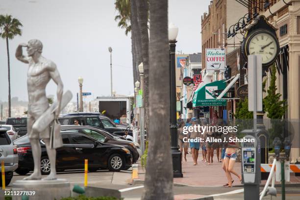 Statue of fallen Newport Beach lifeguard Ben Carlson stands at left as beach-goers walk down the sidewalk past mostly closed businesses on a...
