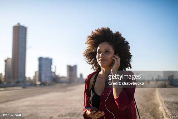 junge lateinamerikanische frau, die musik hört, während sie in der stadt spazieren geht - afro caribbean and american stock-fotos und bilder