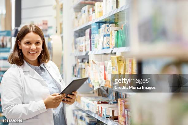 smiling pharmacist holding digital tablet by rack - female pharmacist with a digital tablet imagens e fotografias de stock