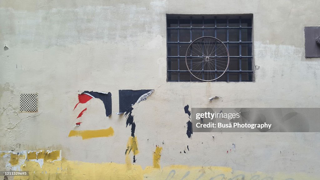 Street wall with a bike wheel on window and scratched posters. In Florence, Italy