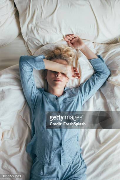 beautiful blonde woman in blue pyjamas lying on bed covering eyes with arm - woman pillow over head stock pictures, royalty-free photos & images