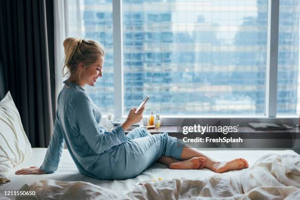 weekend rituals: mooie happy young blonde vrouw in pyjama s sitting comfortabel op bed en met behulp van haar smartphone op een zonnige ochtend - blond women happy eating stockfoto's en -beelden