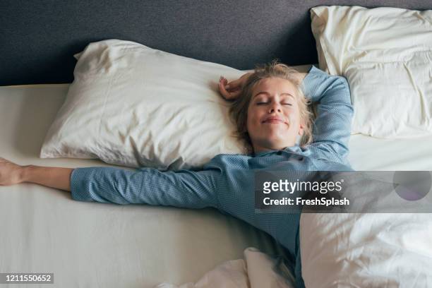 fühlen sie sich belebt: happy blonde woman in pyjamas dehnt sich im bett nach dem aufwachen am morgen - hotel stock-fotos und bilder