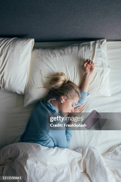 technologie-aided sleep: vrouw in pyjama met draadloze hoofdtelefoon luisteren naar ontspannende muziek op haar smartphone in bed in de ochtend - bed sleep stockfoto's en -beelden