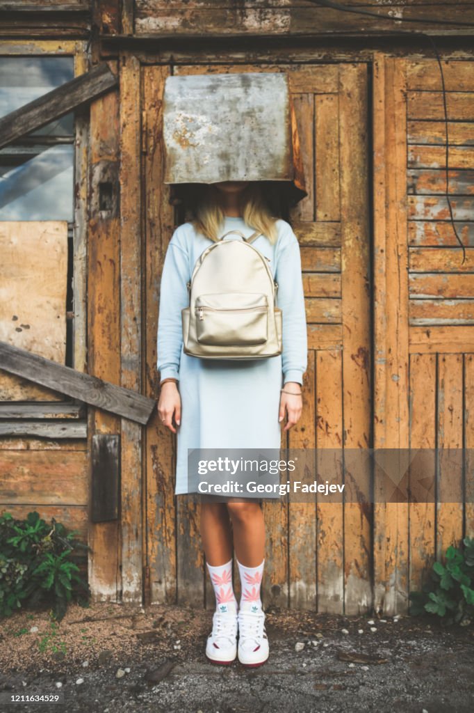 A long haired blonde woman, wearing a powder blue dress, white socks with pink marijuana motifs, white trainers and a pink backpack the wrong way round, wearing on her head a rusty metal container.