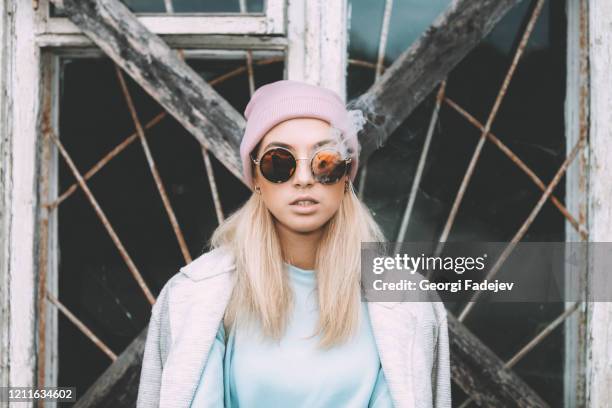 hipster woman smoking a sigarette, wearing a hat and vintage, hipster glasses. a lot of smoke. soft lights. trendy street style. - cannabis narcotic stockfoto's en -beelden