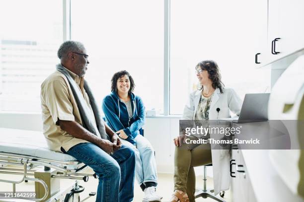 smiling female doctor consulting with senior male patient and adult daughter in exam room - patient room stockfoto's en -beelden