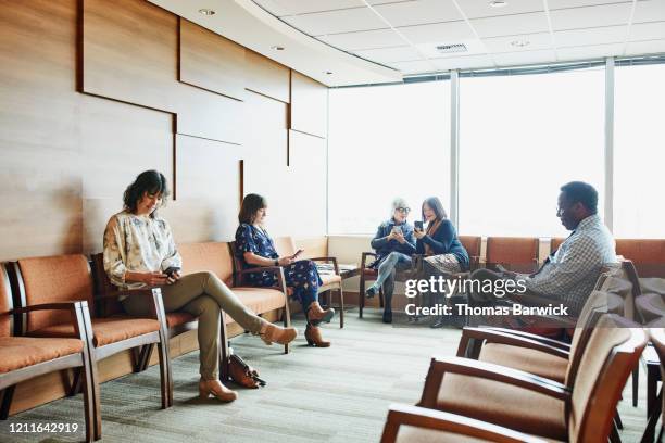 smiling senior women sharing photos on smart phones while sitting in medical office waiting room - waiting room - fotografias e filmes do acervo