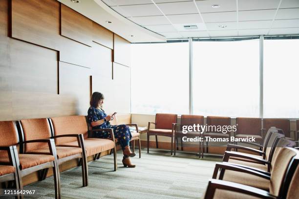 woman looking at smart phone while sitting in medical office waiting room - waiting room - fotografias e filmes do acervo