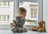 Child in home quarantine at the window putting a medical mask on his sick teddy bear, for protection against viruses during coronavirus COVID-19 and flu outbreak. Children and illness disease concept
