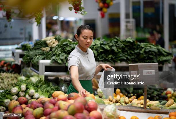 latinamerikansk ung kvinna som arbetar på en frukt- och grönsaksmarknad; stall packning mango i en plastpåse - latin american and hispanic shopping bags bildbanksfoton och bilder