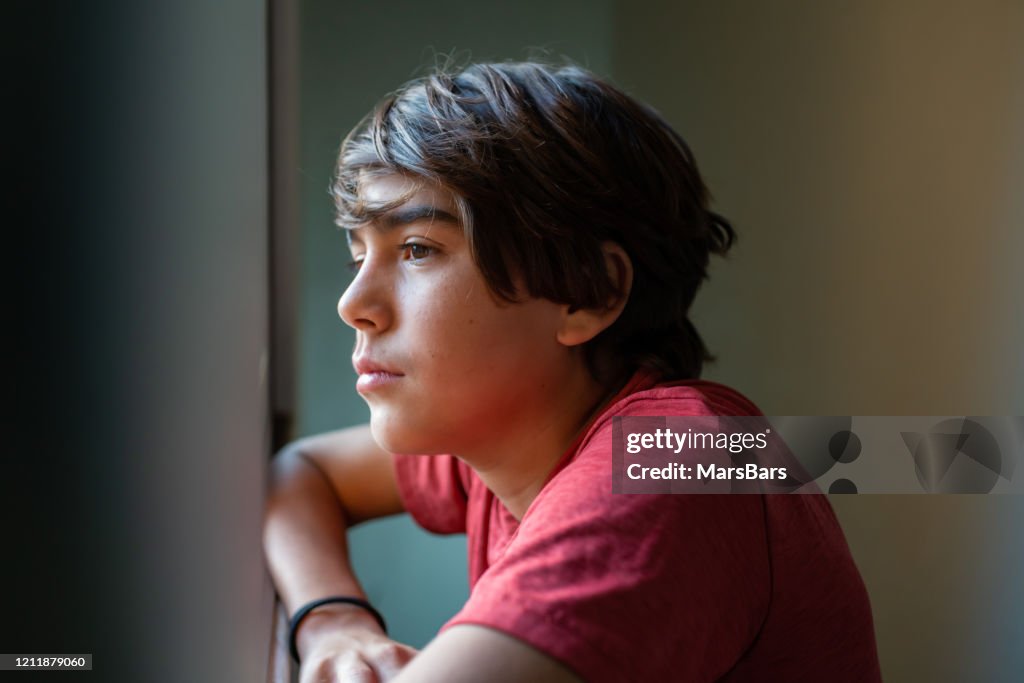 Latinx preadolescent boy looking out through window, reflecting, relaxing, pensive.