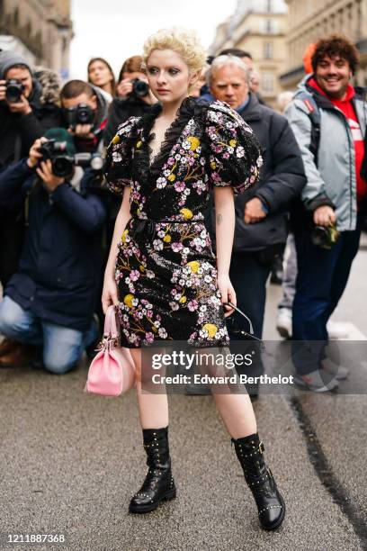 Lachlan Watson wears earrings, a black dress with colorful floral embroideries, puff sleeves and a plunging neckline with a black frilly collar, a...