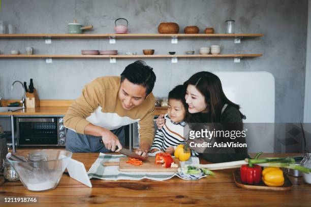 happy young family of three using digital tablet for recipe while preparing food together in the kitchen - asian family cooking stock pictures, royalty-free photos & images