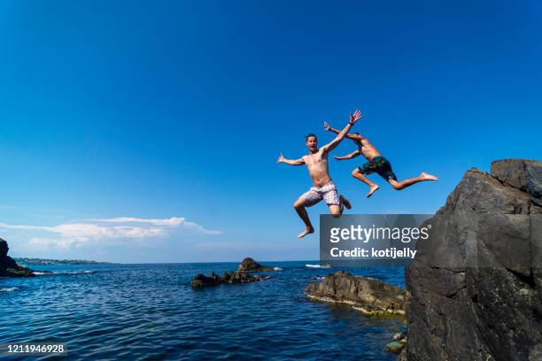 two young men jumping off cliff into sea - cliff dive stock pictures, royalty-free photos & images