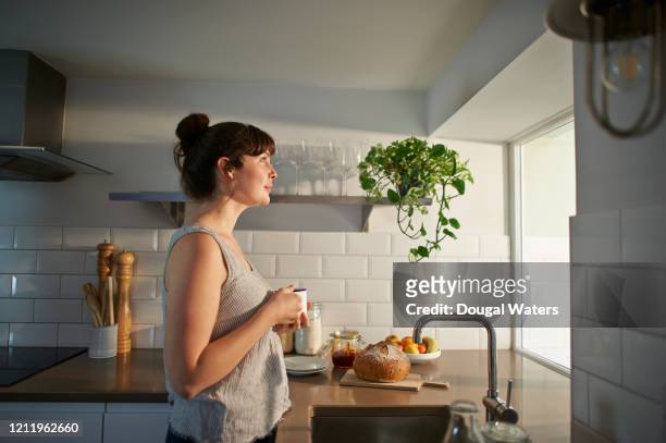 woman holding hot drink and looking out of kitchen window. - day dreaming stock pictures, royalty-free photos & images