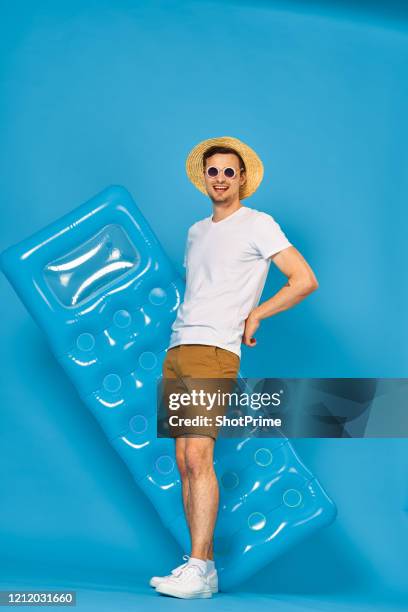 a young man in summer clothes, shorts and a white t-shirt in a straw hat and sunglasses smiles broadly and holds an air mattress in his hands on a blue background. - blue white summer hat background stock pictures, royalty-free photos & images