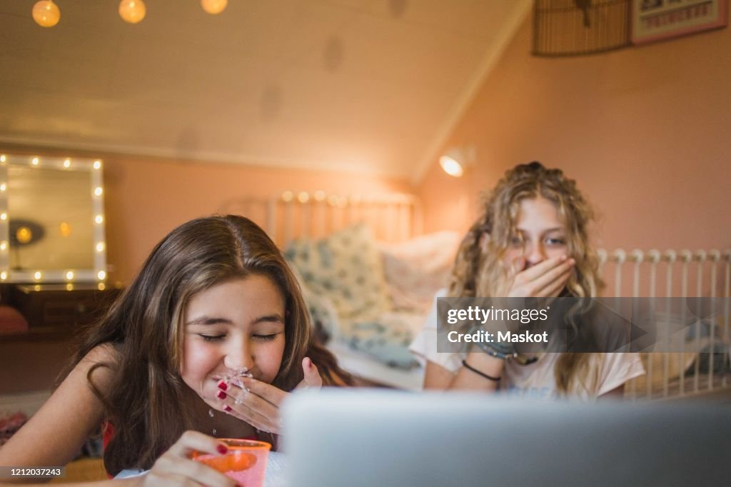 Girl spitting water while playing game and watching laptop with friend in bedroom