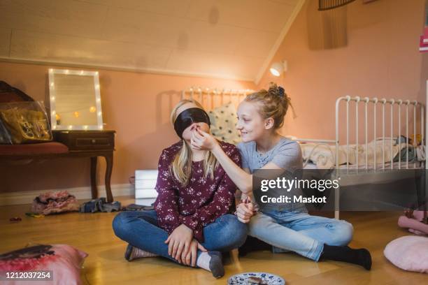 full length of blindfolded girl tasting food while sitting with friend in bedroom - blinddoek stockfoto's en -beelden