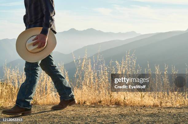 cowboy walking holding a cowboy hat with a mountain range behind him. - cowboy boots stock pictures, royalty-free photos & images