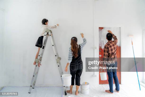 happy asian family with father, mother and daughter with paintbush in hand painting white color a wall together in their new house. family life love relationship, or new house activity concept - couple painting stock pictures, royalty-free photos & images