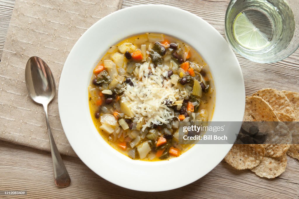 Bowl of vegetable black bean barley soup with grated cheese on a rustic wood table with a spoon, napkin, crackers and a glass of lime infused water