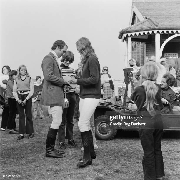 British equestrian Mark Phillips, holding an upturned trophy by the base, with British royal Princess Anne, as a number of children stand around...