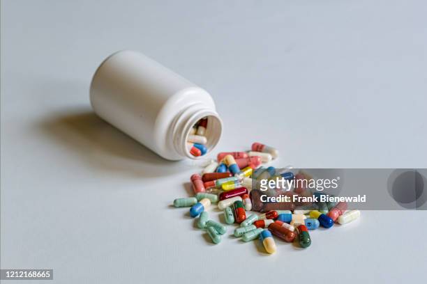 In this photo illustration many differently colored medical capsules, running out of a white container, is displayed on a white table.