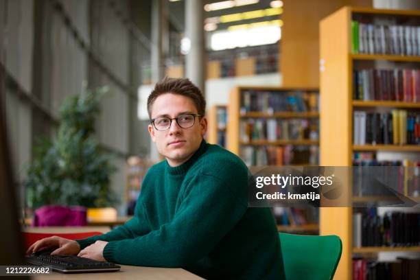 portrait of young adult man sitting in library by a computer and looking around - northern european descent stock pictures, royalty-free photos & images