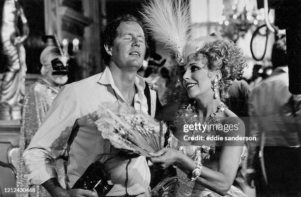 Actress Joan Collins uses her fan to cool down photographer Eddie Sanderson during the Golden Ball on the set of the miniseries Sins, filmed in...