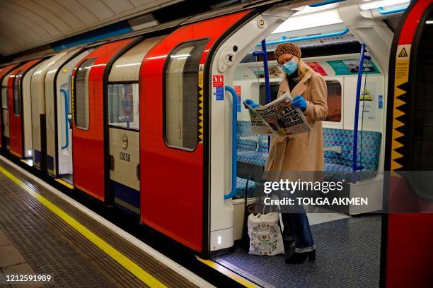 Woman wearing PPE , including a face mask as a precautionary measure against COVID-19, reads a newspaper as she stands aboard a London Underground...