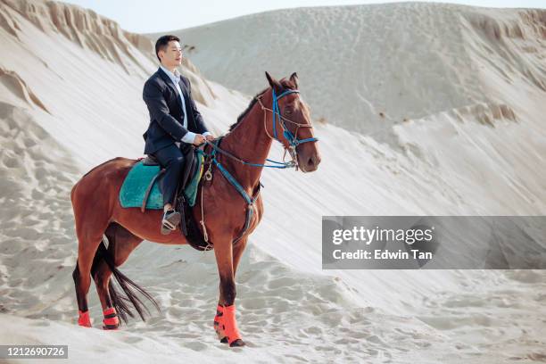 ein asiatisches chinesisches männchen mit formeller kleidung reiten ein pferd in der wüste sanddüne von melaka während des sonnenuntergangs - horse and male and riding stock-fotos und bilder