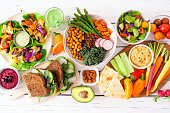 Healthy lunch table scene with nutritious lettuce wraps, Buddha bowl, vegetables, sandwiches, and salad, overhead view over white wood