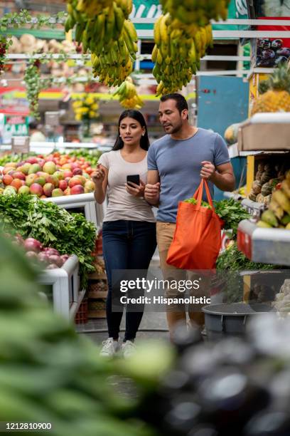 young couple buying fruits and vegetables while woman looks at shopping list on smartphone's app - fruit stand stock pictures, royalty-free photos & images