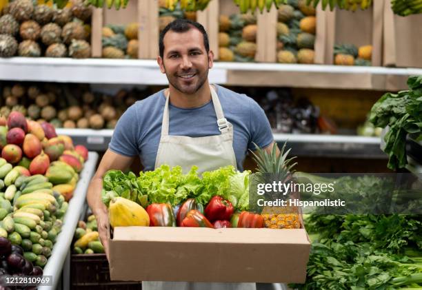 porträt eines hübschen arbeiters auf einem bauernmarkt, der produkte im karton hält, während er lächelnd mit der kamera zu sieht - marktplatz stock-fotos und bilder