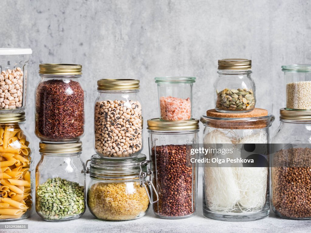 Glazen potten met verschillende granen en zaden - erwten split, zonnebloem- en pompoenpitten, bonen, rijst, pasta, havermout, linzen, bulgur op een grijze achtergrond