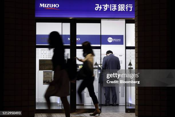 Pedestrians walk past a customer using an automated teller machine at a branch of Mizuho Bank Ltd., a unit of Mizuho Financial Group Inc. , at night...