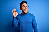 Young handsome man with beard wearing casual sweater and glasses over blue background Waiving saying hello happy and smiling, friendly welcome gesture