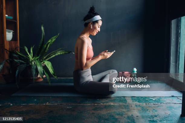 elegir música de entrenamiento: mujer asiática con auriculares sentadoen en la alfombra de ejercicio y configurar una lista de reproducción en su smarphone - entrenamiento en casa fotografías e imágenes de stock