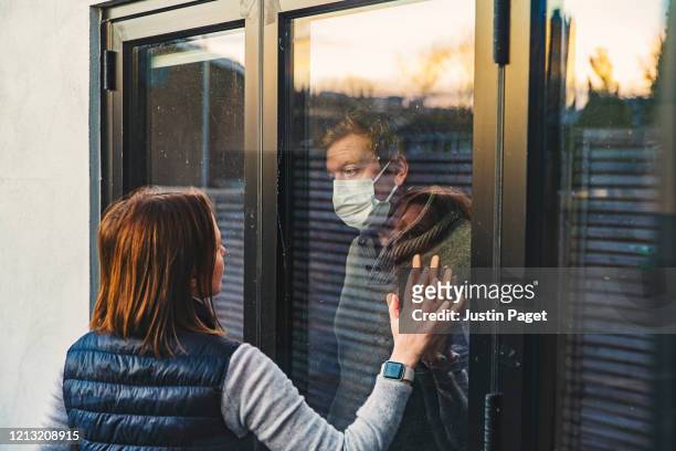 woman looking at masked husband quarantined behind window - quarantäne stock-fotos und bilder