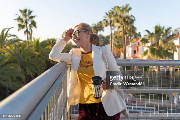Senior woman standing on bridge, enjoying the sunshine, holding reuseable cup